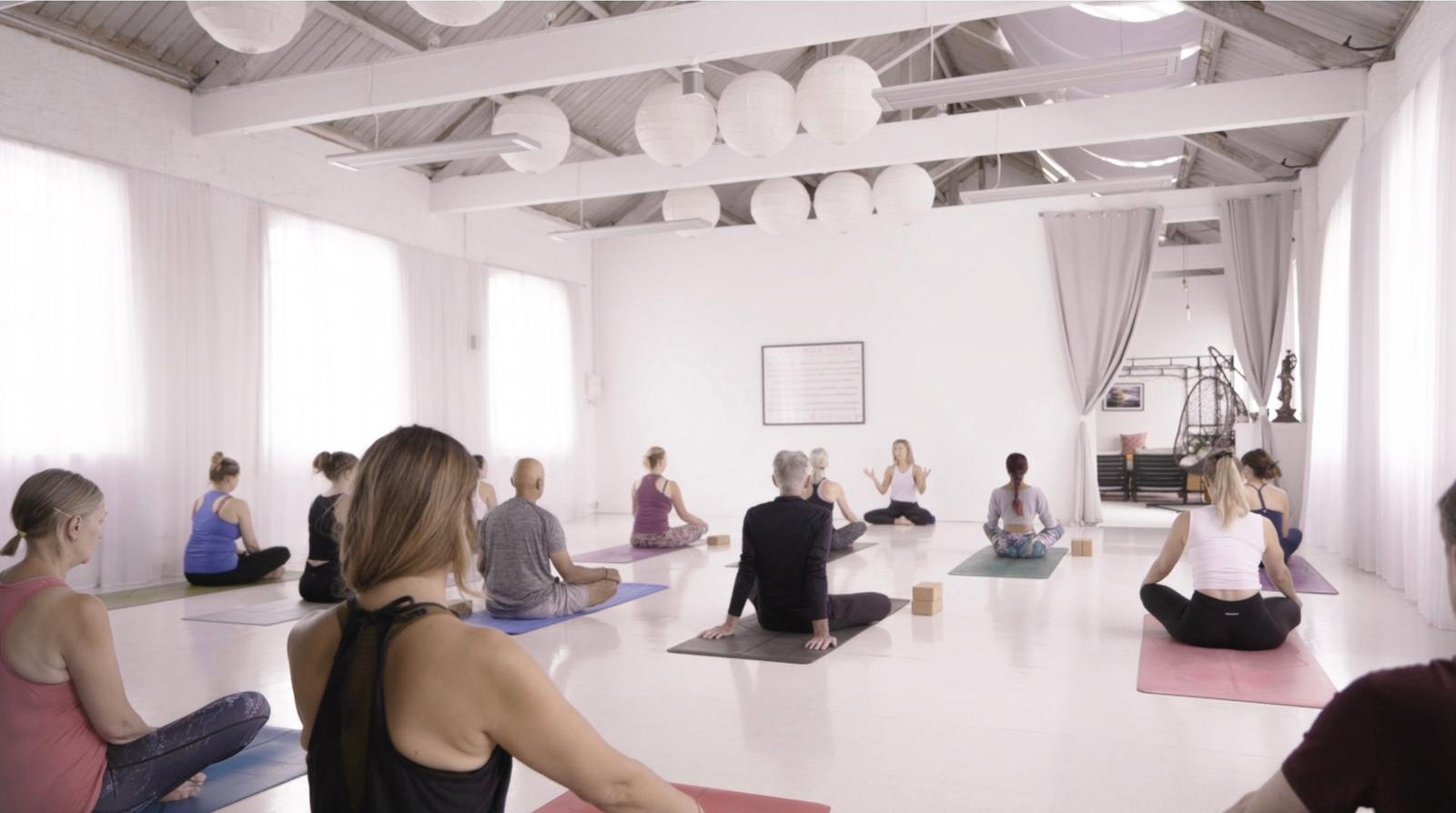 A class sitting cross-legged on their mats while the teacher speaks at the front of the studio